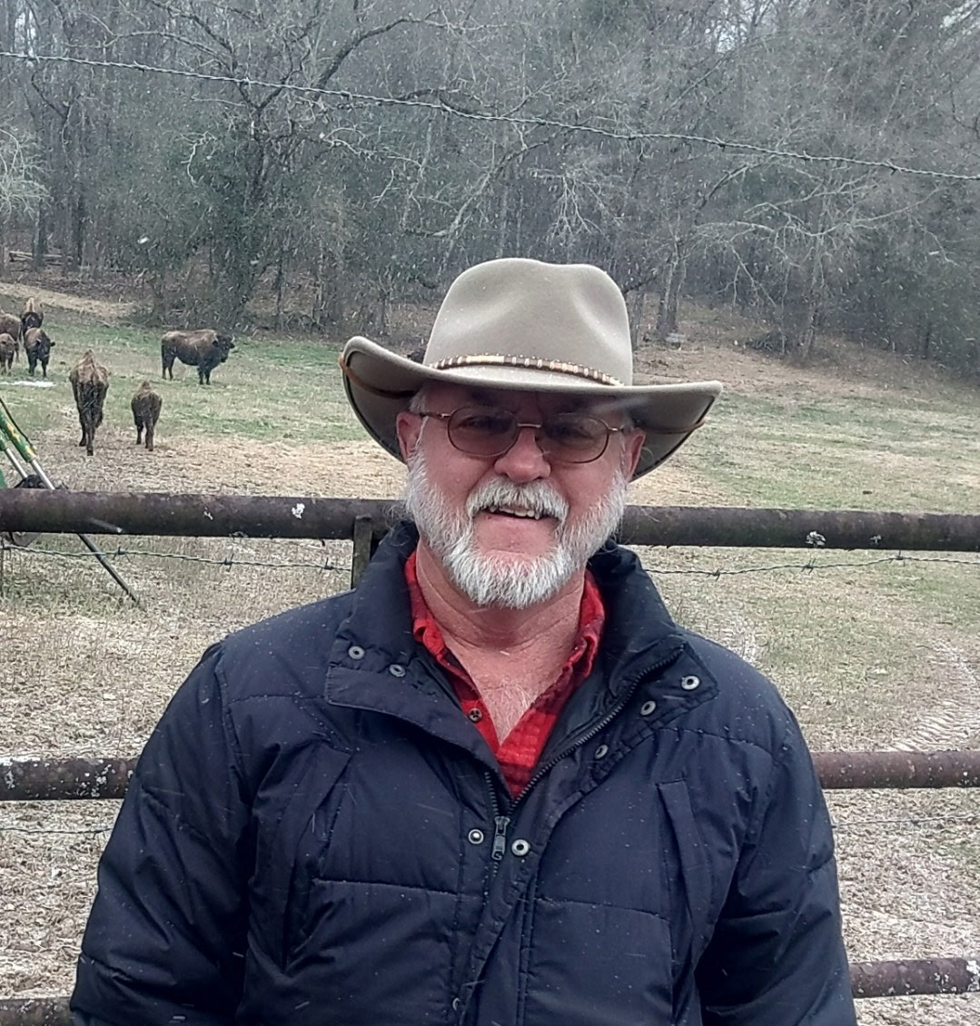 DJ White at the fence with bison grazing in the pasture behind him