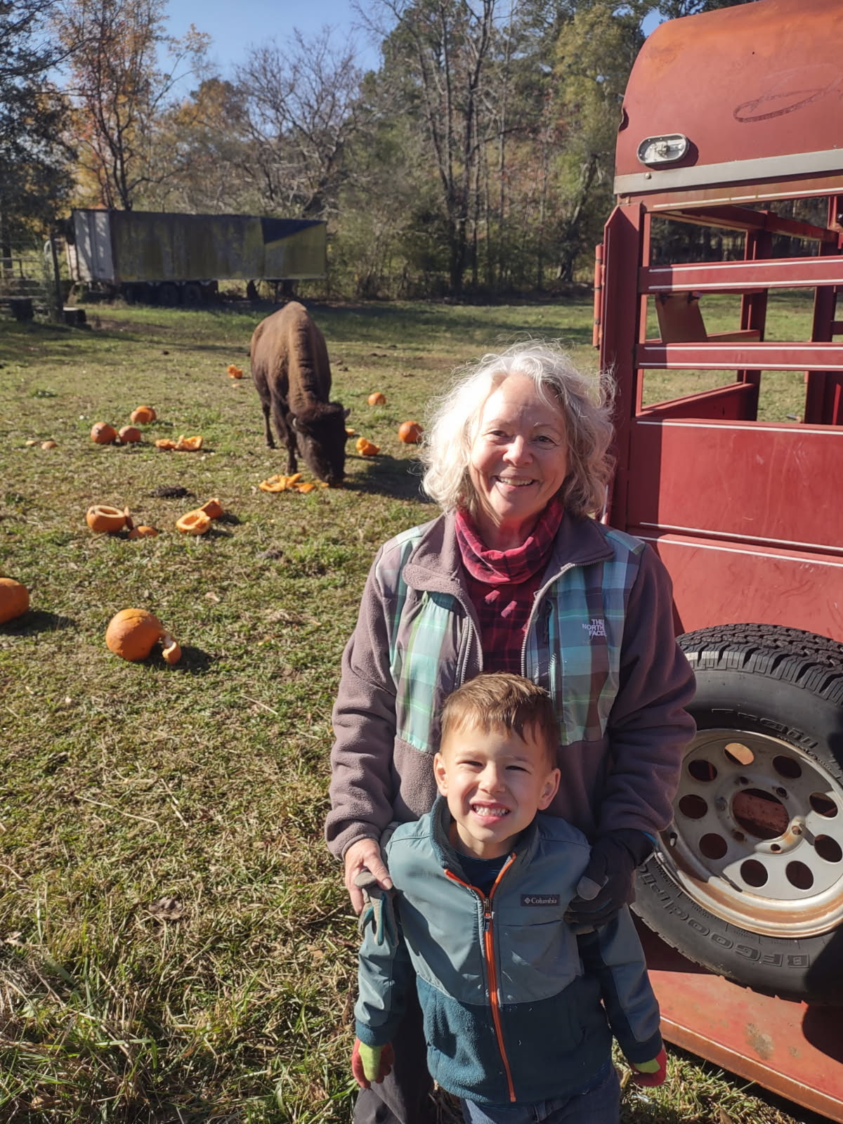 Mischa White with a grandson at the ranch, a bison grazing on pumpkins in the pasture behind them